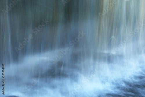 Water flows down a small wall with reflections behind. Blue-white blurred water background. Blurred artificial waterfall background.