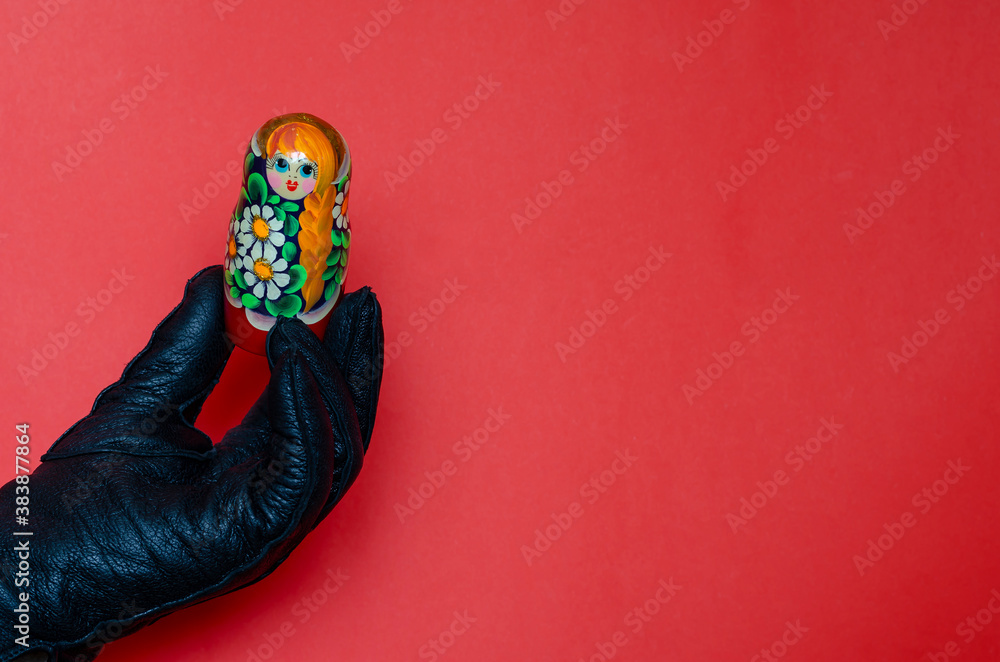 A hand in a black leather glove holds a matryoshka doll on a red background.