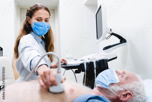 Young woman doctor in mask checking up elderly patient chest with ultrasound equipment photo