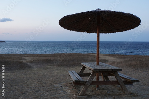 Wooden bench with table and straw umbrella without people on a high beach with sea view at sunset © Anton