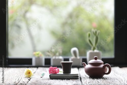 tea cup with clay teapot and notebooks with flowers on old brown table with cactus plant in front of windows 