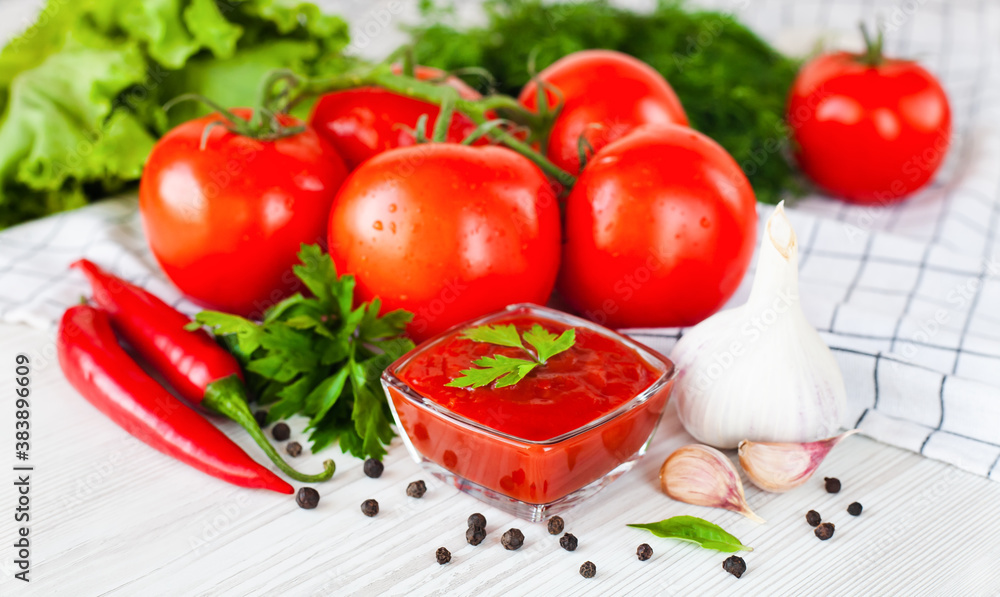 Red sauce or ketchup in a glass bowl and ingredients for his cooking, tomatoes, garlic, basil, parsley, red hot pepper and spices on a wooden white background. Close-up.