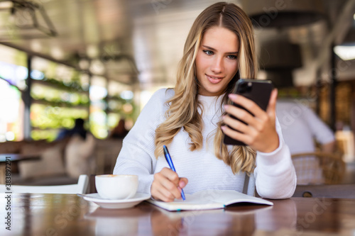 Young woman reading news from social networks using mobile phone at cafe. Female blogger making online research on smartphone while creating article for share writing in notepad