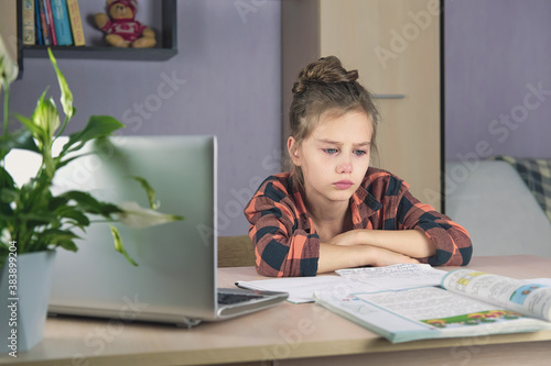 A girl sits at the table at home, does homework with a laptop and cries