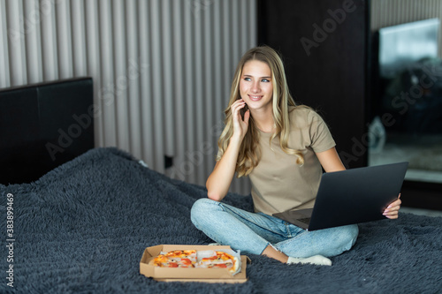 Young woman use laptop and ear pizza from box sitting on a bed in bedroom. Food Delivery concepr photo