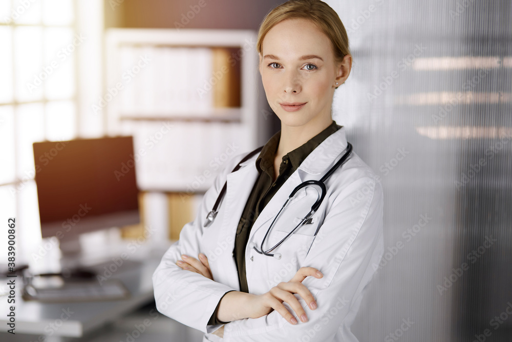 Cheerful smiling female doctor standing in sunny clinic. Portrait of friendly physician woman. Perfect medical service in hospital. Medicine concept