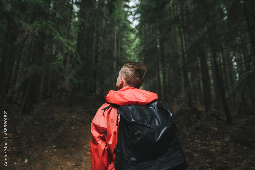 Back view of a young male hiker with a travel backpack wearing a red raincoat looking at the mountain forest. Rainy weather, hiking concept.
