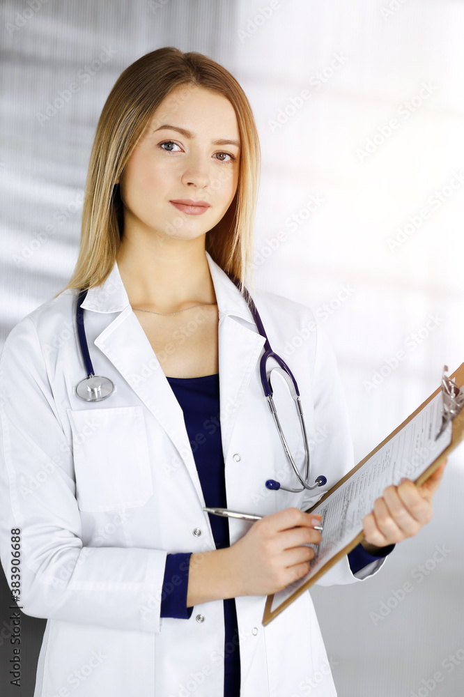 Young optimistic woman-doctor is holding a clipboard in her hands, while standing in a sunny cabinet of a clinic. Portrait of friendly female physician with a stethoscope. Perfect medical service in a Young optimistic woman-doctor is holding a clipboard in her hands, while standing in a sunny cabinet of a clinic. Portrait of friendly female physician with a stethoscope. Perfect medical service in a