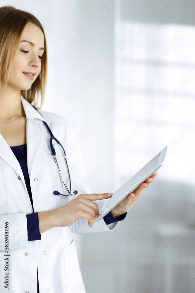 Young woman-doctor is holding a tablet computer in her hands, while standing in a clinic. Portrait of friendly female physician with a stethoscope at work. Perfect medical service in a hospital Young woman-doctor is holding a tablet computer in her hands, while standing in a clinic. Portrait of friendly female physician with a stethoscope at work. Perfect medical service in a hospital