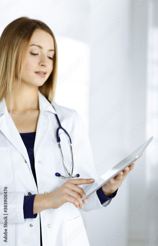 Young woman-doctor is holding a tablet computer in her hands, while standing in a clinic. Portrait of friendly female physician with a stethoscope at work. Perfect medical service in a hospital Young woman-doctor is holding a tablet computer in her hands, while standing in a clinic. Portrait of friendly female physician with a stethoscope at work. Perfect medical service in a hospital