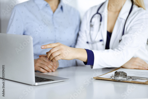 Unknown woman- doctor and her patient are discussing patient s blood test  while sitting together at the desk in the cabinet in a clinic. Female physician  with a stethoscope  is using a laptop