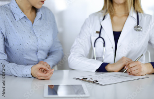 Unknown woman- doctor is listening to her patient, while sitting together at the desk in the cabinet in a clinic. Female physician with a stethoscope is writing at clipboard, close up. Perfect medical