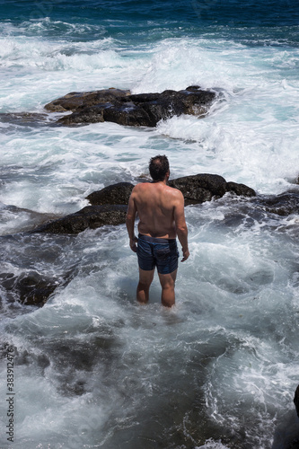 An unrecognizable sturdy man in blue trunks stands in the sea at daytime, facing the challenging strong currents and surf that hit the rocks of the coast.