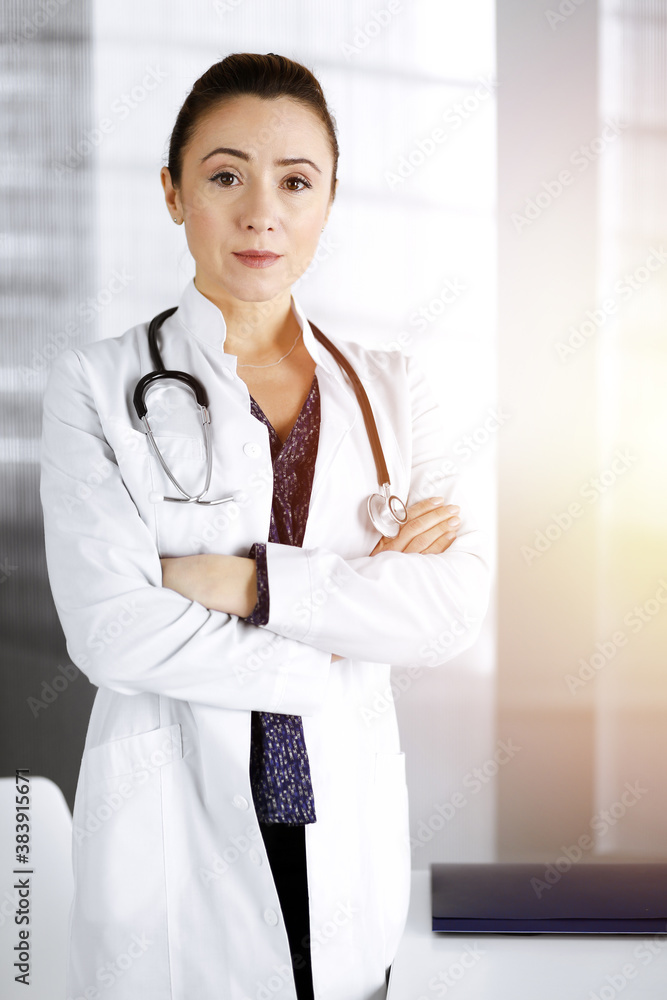 Smiling professional woman-doctor is standing with crossed arms in a sunny clinic office. Portrait of friendly female physician with a stethoscope at work. Perfect medical service in a hospital Smiling professional woman-doctor is standing with crossed arms in a sunny clinic office. Portrait of friendly female physician with a stethoscope at work. Perfect medical service in a hospital