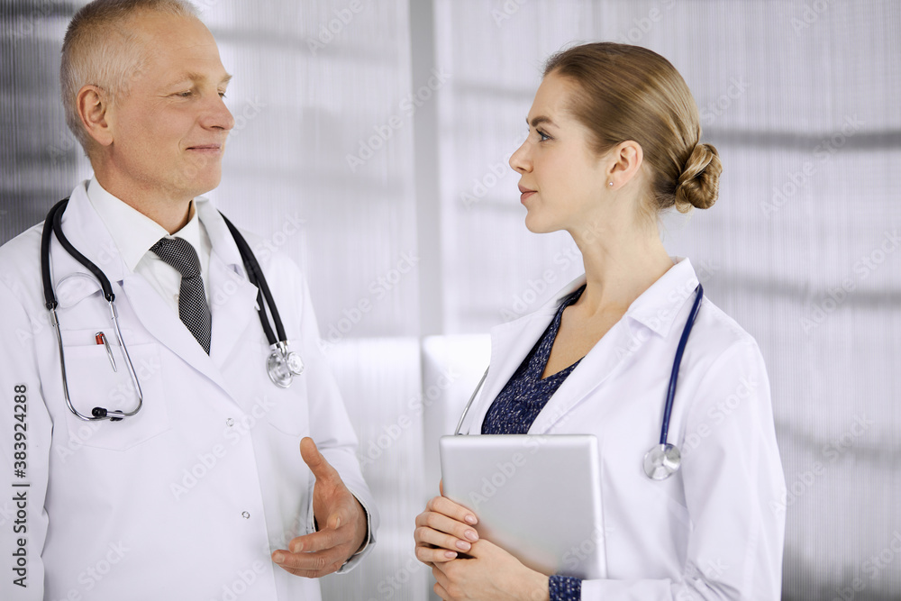 Elderly confident doctor and woman physician discussing current disease therapy while standing in clinic. Teamwork in hospital. Medicine and healthcare concept Elderly confident doctor and woman physician discussing current disease therapy while standing in clinic. Teamwork in hospital. Medicine and healthcare concept
