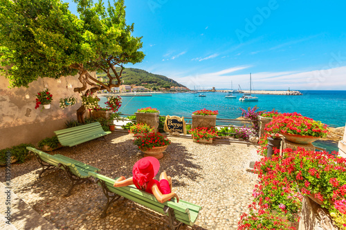 Lifestyle woman in red sitting on bench in Marciana Marina, flowery old district Borgo al Cotone: meaning COTTON VILLAGE, looking harbor bay with ships. Tourist on holiday travel in Italy, Elba island