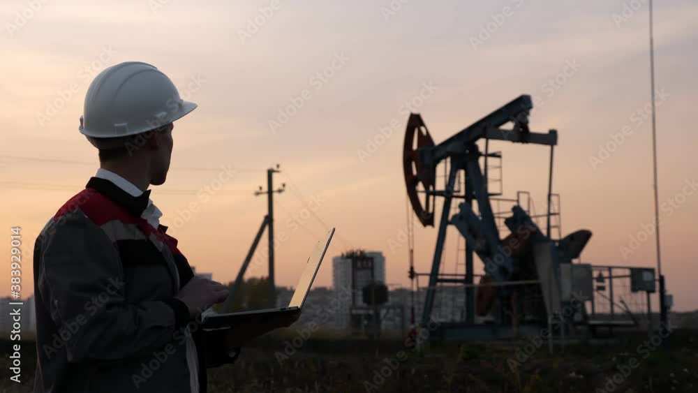 man in a white helmet with a laptop on the background of an oil rocking at sunset. man in a white helmet with a laptop on the background of an oil rocking at sunset.
