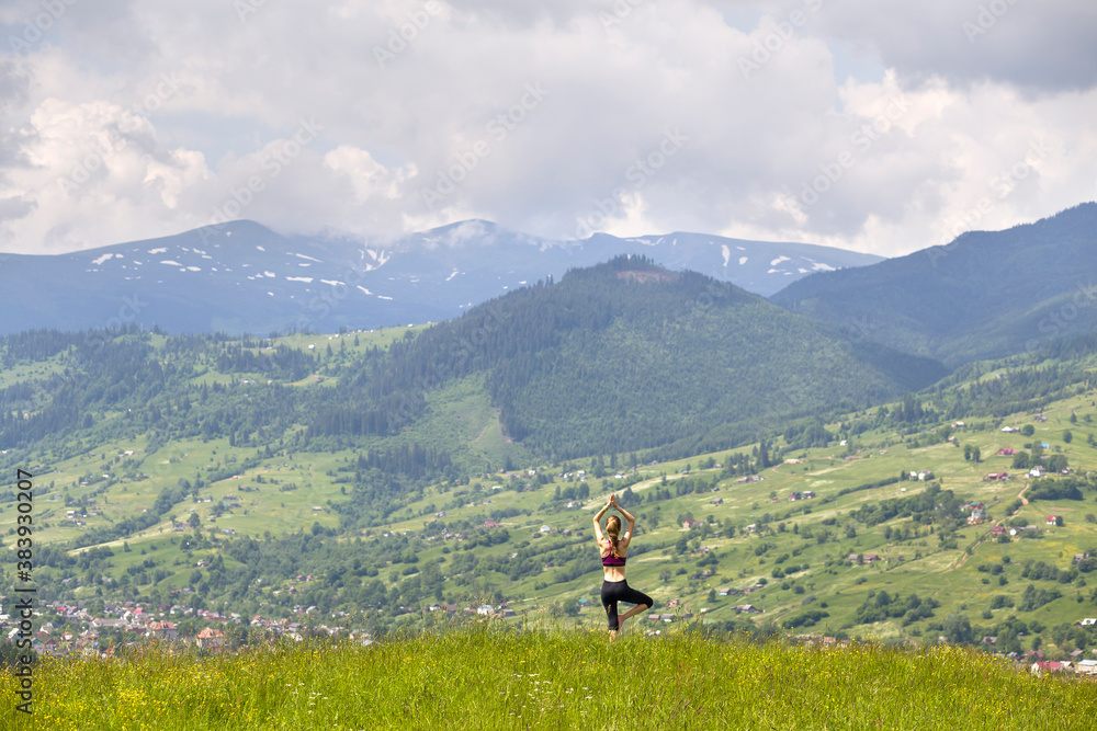 Attractive slim young woman doing yoga exercises outdoors on background of green mountains on sunny summer day.