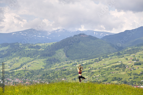 Attractive slim young woman doing yoga exercises outdoors on background of green mountains on sunny summer day.