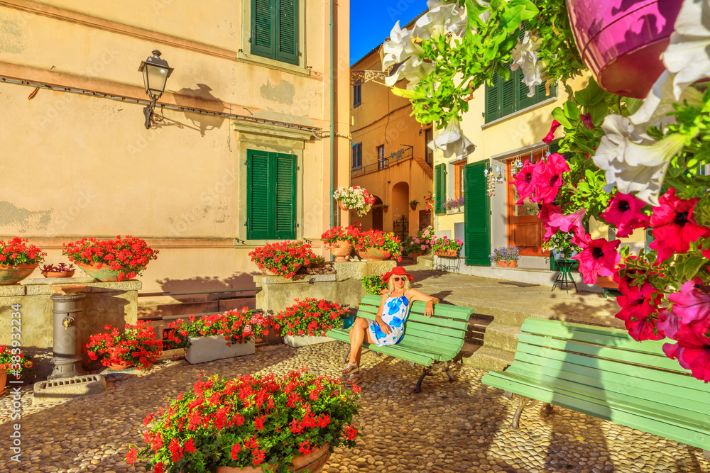 Woman sitting on bench in Marciana Marina, flowery old district Borgo al Cotone: meaning COTTON VILLAGE. Tourist travel in Italy. Marciana Marina, Elba island. Blooms of red geraniums on foreground.