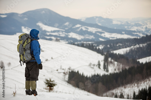 Back view of tourist hiker in warm clothing with backpack standing on mountain clearing on copy space background of woody mountain ridge and cloudy sky.