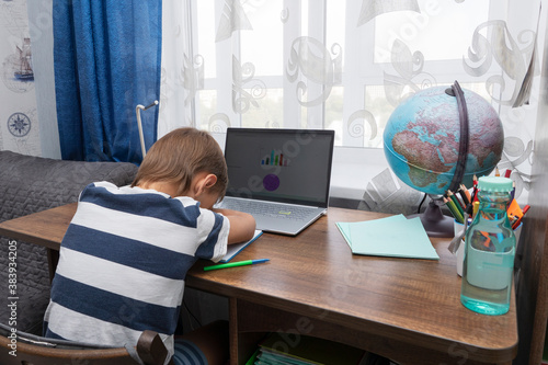 schoolboy is sleeping on desk by laptop while doing homework on distant learning.. Home education concept. Students are exhausted doing excess homework.