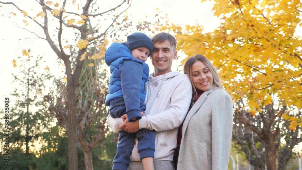 Lovely family of mother father and funny little boy hug posing for camera in picturesque autumn city park on nice warm day low angle shot Lovely family of mother father and funny little boy hug posing for camera in picturesque autumn city park on nice warm day low angle shot