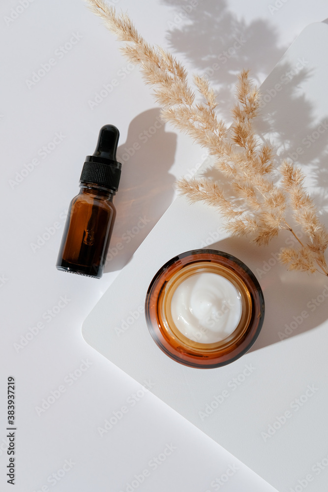 Set of eco natural cosmetics on white podium. Amber glass dropper bottle with essential oil and jar of moisturizer, dried flowers. View from above.