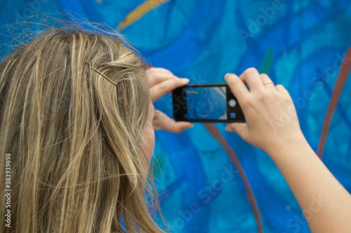 Mujer rubia sujeta con dos manos móbil negro y saca foto al muro azul del fofo