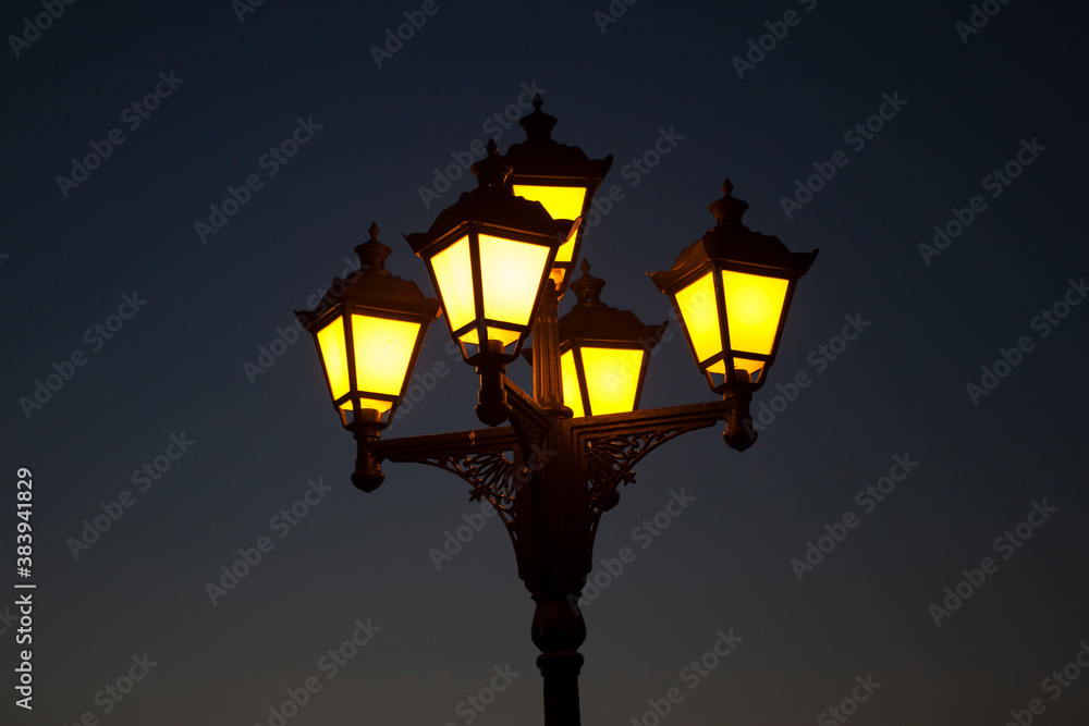 Street city cast-iron lantern lit at night, yellow lantern light on dark blue background.