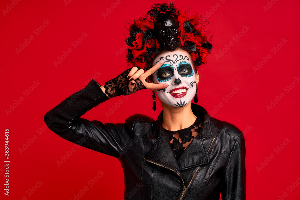Dead gir with sugar skull makeup with a wreath of flowers on her head and skull, wearth lace gloves and showing victory sign isolated on red background.Concept of Halloween or La Calavera Catrina