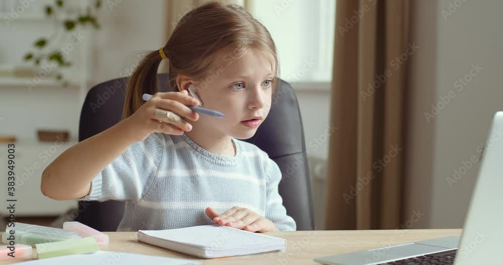 Portrait of little cute preschool girl wears headphones sits at home table alone looks at laptop screen repeats movements of online teacher draws on paper with pencil pen, concept of remote e-learning Portrait of little cute preschool girl wears headphones sits at home table alone looks at laptop screen repeats movements of online teacher draws on paper with pencil pen, concept of remote e-learning