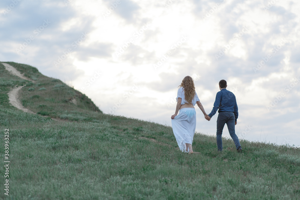 A couple in love, a pregnant wife in a white outfit are walking in a hilly field. Overcast weather A couple in love, a pregnant wife in a white outfit are walking in a hilly field. Overcast weather