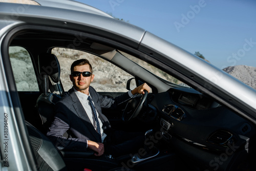 man businessman in a jacket, shirt, tie and sunglasses is driving a car.
