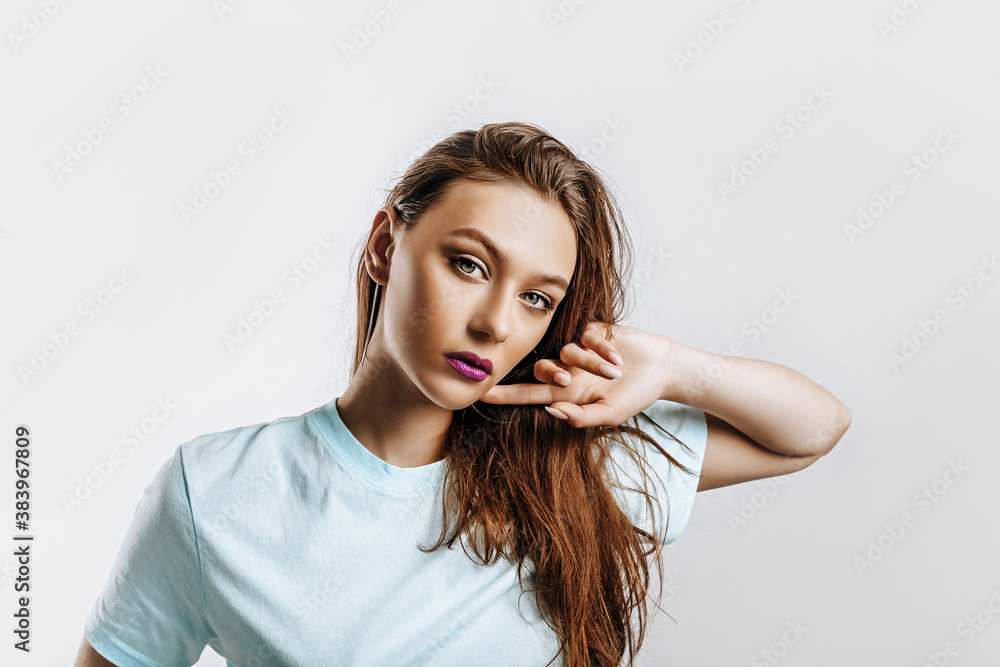 Beautiful young brunette girl with green eyes with long hair with fashion makeup with purple lips on an isolated gray background. The woman holds hand to face and seriously looking at the camera. Beautiful young brunette girl with green eyes with long hair with fashion makeup with purple lips on an isolated gray background. The woman holds hand to face and seriously looking at the camera.