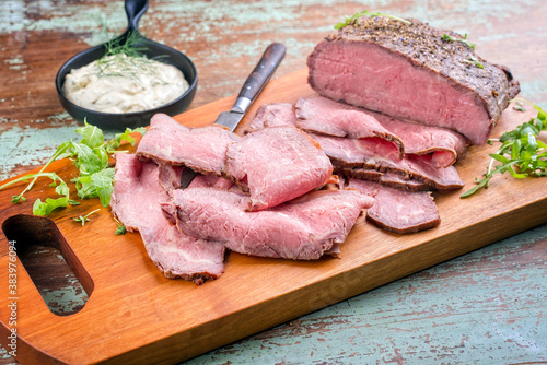 Modern style traditional lunch meat with sliced cold cuts roast beef with rocket salad and remoulade offered as top view on a wooden design board