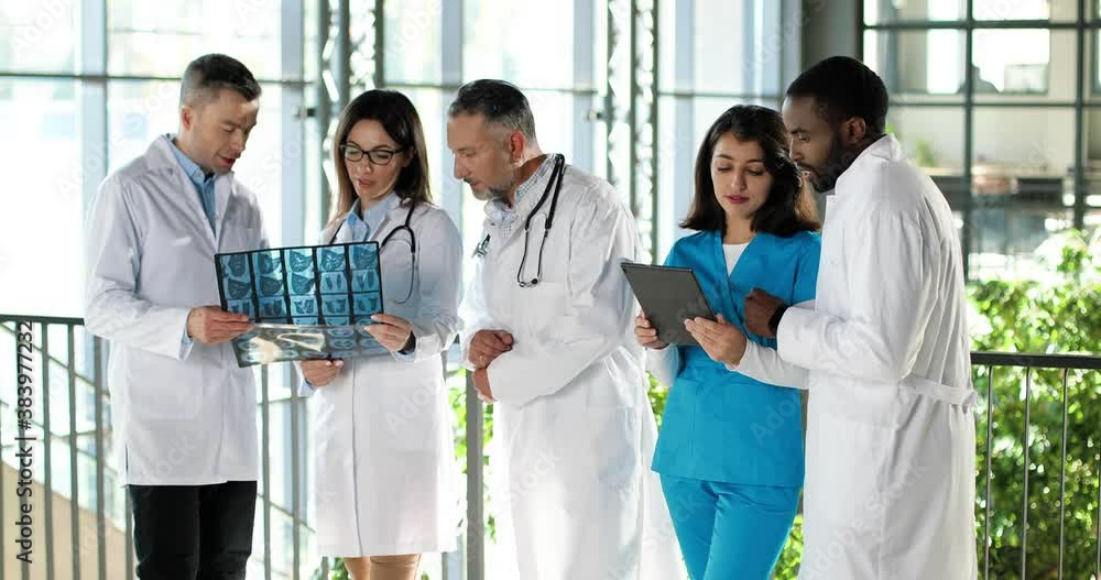 Multi ethnic doctors, men and women, standing in clinic, talking and discussing diagnosis. Mixed-races males and females physicians having discussion in hospital with X-ray and tablet device. Multi ethnic doctors, men and women, standing in clinic, talking and discussing diagnosis. Mixed-races males and females physicians having discussion in hospital with X-ray and tablet device.