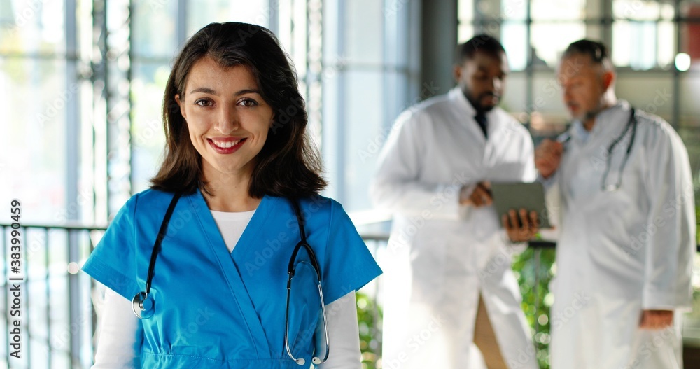 Portrait of Caucasian happy beautiful woman physician smiling to camera and holding documents in clinic. Indoor. Pretty female doctor in hospital. Multi ethnic doctors talking on background. Portrait of Caucasian happy beautiful woman physician smiling to camera and holding documents in clinic. Indoor. Pretty female doctor in hospital. Multi ethnic doctors talking on background.