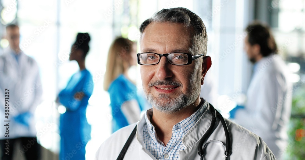 Fototapeta premium Portrait of Caucasian senior man physician in white gown, stethoscope and glasses looking at camera and smiling in clinic. Handsome male doctor in hospital. Doctors on background. Healthcare.
