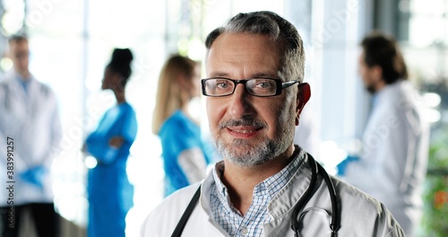 Portrait of Caucasian senior man physician in white gown, stethoscope and glasses looking at camera and smiling in clinic. Handsome male doctor in hospital. Doctors on background. Healthcare.