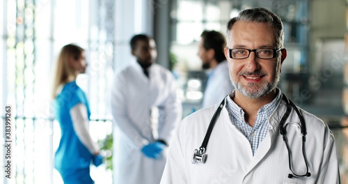 Portrait shot of Caucasian senior man physician in white gown, stethoscope and glasses looking at camera and smiling in clinic. Handsome male doctor in hospital. Doctors on background Medicine concept