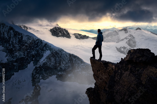 Man Hiker on top of a Mountain Peak. Dreamscape Artistic Render Composite. Landscape background from British Columbia, Canada. Dark Dramatic Sunset Sky.