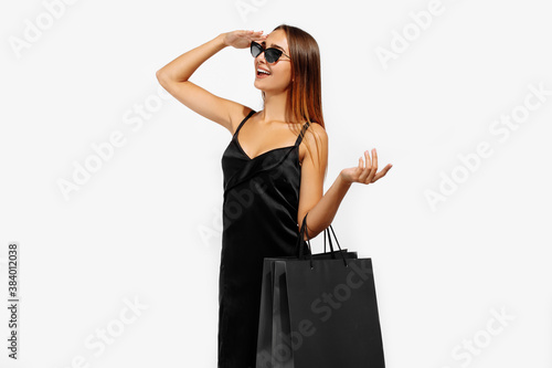 Beautiful young woman in sunglasses, in a black dress holds bags and looks into the distance on a white background, Black Friday, shopping