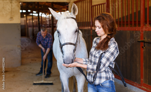 Female farmer standing with horse, man cleaning floor at stabling