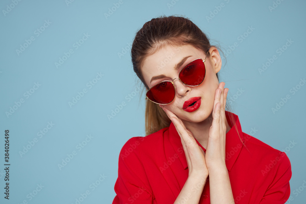 Woman in red shirt on blue background holds hands near face and sunglasses red lips makeup model Woman in red shirt on blue background holds hands near face and sunglasses red lips makeup model