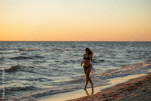 A young beautiful woman in a black bikini and a white shirt on a tanned body is walking along the beach at sunset. Soft selective focus, art nose.