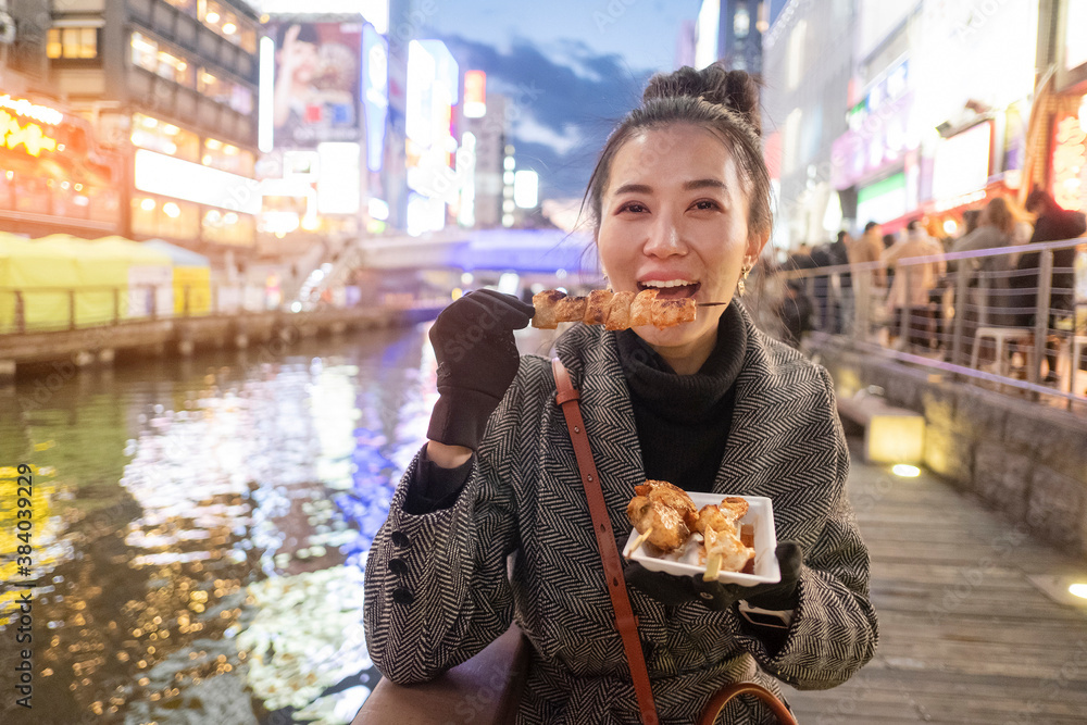 Fototapeta premium Young woman tourists enjoy eating street food in walking at street shopping center Dotonbori in Osaka, Japan.