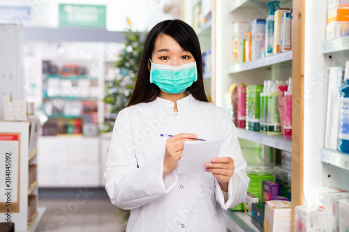 Diligent friendly smiling chinese woman pharmacist in protective facial mask keeps track of drugs in interior of pharmacy