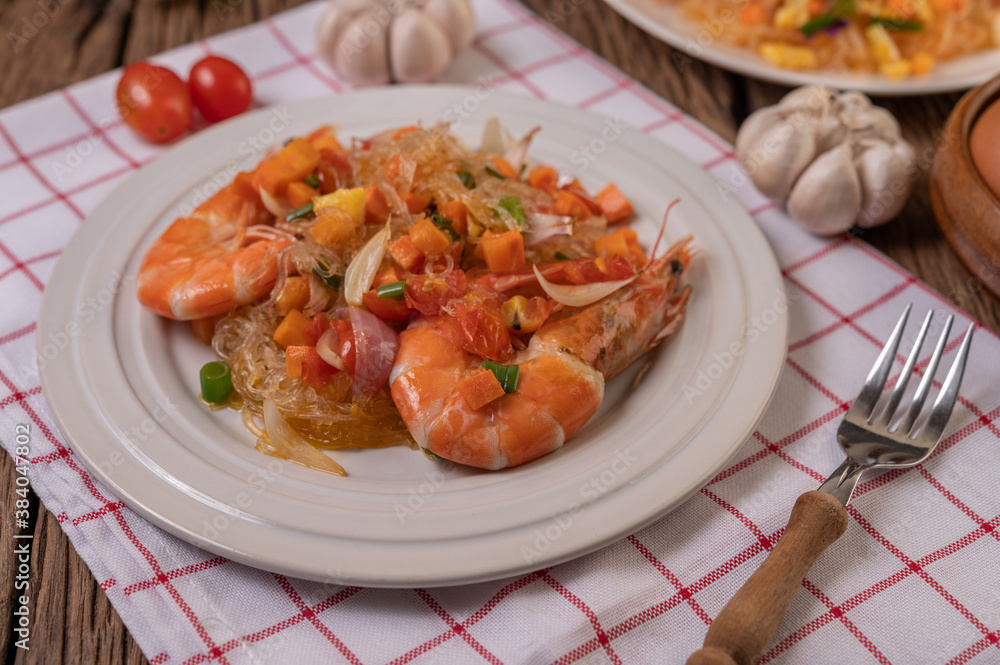 Stir-fried prawns with glass noodles in a white plate placed on a cloth with eggs and garlic. Stir-fried prawns with glass noodles in a white plate placed on a cloth with eggs and garlic.