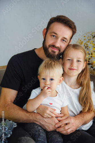 Portrait of a happy father with adorable children sitting on the sofa posing for a family photo at a holiday home. Cuddles with cute little son and daughter while looking at the camera © Elena 